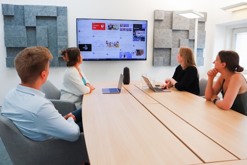 Four people are seated around a conference table in a modern office setting. A large screen on the wall displays various content, suggesting a presentation or discussion. The room is well-lit, with acoustic panels on the walls, and two laptops are open on the table.
