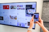 A person uses a smartphone to scan a QR code displayed on a large screen mounted on a wall. The screen shows various social media posts and images, including photos of people and text content. The hands of the person holding the phone are visible, and the setting appears to be indoors with a subtle stone wall design on the right.