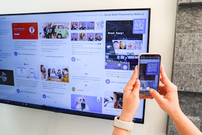 A person uses a smartphone to scan a QR code displayed on a large screen mounted on a wall. The screen shows various social media posts and images, including photos of people and text content. The hands of the person holding the phone are visible, and the setting appears to be indoors with a subtle stone wall design on the right.