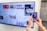 A person uses a smartphone to scan a QR code displayed on a large screen mounted on a wall. The screen shows various social media posts and images, including photos of people and text content. The hands of the person holding the phone are visible, and the setting appears to be indoors with a subtle stone wall design on the right.