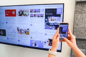 A person uses a smartphone to scan a QR code displayed on a large screen mounted on a wall. The screen shows various social media posts and images, including photos of people and text content. The hands of the person holding the phone are visible, and the setting appears to be indoors with a subtle stone wall design on the right.