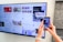 A person uses a smartphone to scan a QR code displayed on a large screen mounted on a wall. The screen shows various social media posts and images, including photos of people and text content. The hands of the person holding the phone are visible, and the setting appears to be indoors with a subtle stone wall design on the right.