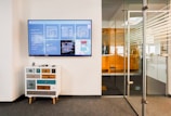 A modern office space features a wall-mounted digital screen displaying various content and information. Below the screen, there is a small, eclectic chest of drawers with a mix of colorful panels. To the right, a glass door with horizontal striped patterns leads to another office area.