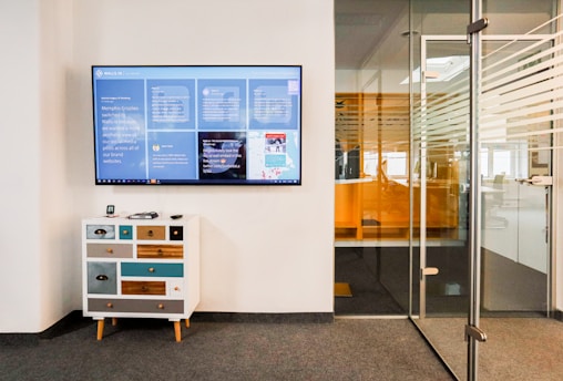 A modern office space features a wall-mounted digital screen displaying various content and information. Below the screen, there is a small, eclectic chest of drawers with a mix of colorful panels. To the right, a glass door with horizontal striped patterns leads to another office area.