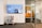 A modern office space features a wall-mounted digital screen displaying various content and information. Below the screen, there is a small, eclectic chest of drawers with a mix of colorful panels. To the right, a glass door with horizontal striped patterns leads to another office area.