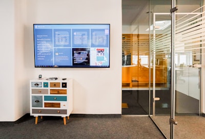 A modern office space features a wall-mounted digital screen displaying various content and information. Below the screen, there is a small, eclectic chest of drawers with a mix of colorful panels. To the right, a glass door with horizontal striped patterns leads to another office area.