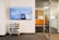 A modern office space features a wall-mounted digital screen displaying various content and information. Below the screen, there is a small, eclectic chest of drawers with a mix of colorful panels. To the right, a glass door with horizontal striped patterns leads to another office area.