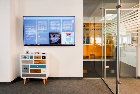 A modern office space features a wall-mounted digital screen displaying various content and information. Below the screen, there is a small, eclectic chest of drawers with a mix of colorful panels. To the right, a glass door with horizontal striped patterns leads to another office area.