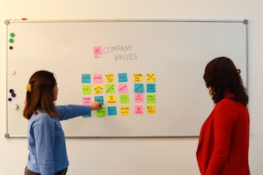 Two people are standing in front of a whiteboard containing various colorful sticky notes. The notes are arranged in a grid and each one has a word or phrase related to company values, such as 'Culture', 'Vision', 'Diversity', and 'Respect'. One person is gesturing towards the board, wearing a blue sweater, while the other is observing attentively, dressed in a red jacket.