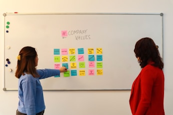 Two people are standing in front of a whiteboard containing various colorful sticky notes. The notes are arranged in a grid and each one has a word or phrase related to company values, such as 'Culture', 'Vision', 'Diversity', and 'Respect'. One person is gesturing towards the board, wearing a blue sweater, while the other is observing attentively, dressed in a red jacket.