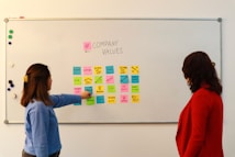 Two people are standing in front of a whiteboard containing various colorful sticky notes. The notes are arranged in a grid and each one has a word or phrase related to company values, such as 'Culture', 'Vision', 'Diversity', and 'Respect'. One person is gesturing towards the board, wearing a blue sweater, while the other is observing attentively, dressed in a red jacket.