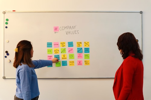 Two people are standing in front of a whiteboard containing various colorful sticky notes. The notes are arranged in a grid and each one has a word or phrase related to company values, such as 'Culture', 'Vision', 'Diversity', and 'Respect'. One person is gesturing towards the board, wearing a blue sweater, while the other is observing attentively, dressed in a red jacket.