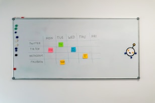 A whiteboard with a weekly social media content schedule is displayed, featuring days of the week across the top and various social media platforms listed on the side. Colorful sticky notes indicate content themes like 'culture', 'fun fact', 'product', and 'how-to'. There are magnets on the left and an eye-catching illustration on the right.