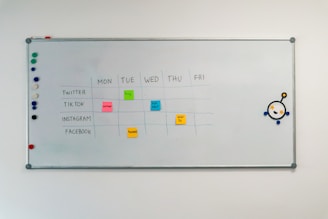 A whiteboard with a weekly social media content schedule is displayed, featuring days of the week across the top and various social media platforms listed on the side. Colorful sticky notes indicate content themes like 'culture', 'fun fact', 'product', and 'how-to'. There are magnets on the left and an eye-catching illustration on the right.