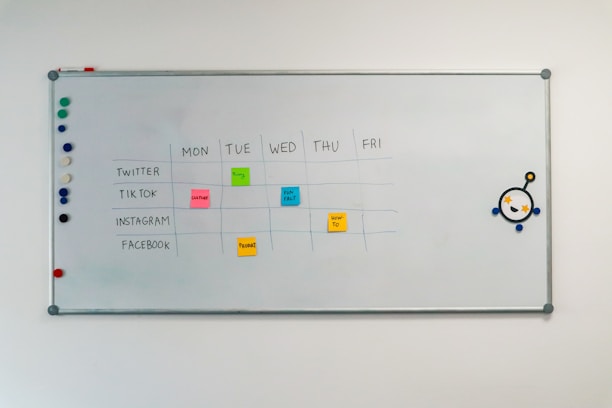 A whiteboard with a weekly social media content schedule is displayed, featuring days of the week across the top and various social media platforms listed on the side. Colorful sticky notes indicate content themes like 'culture', 'fun fact', 'product', and 'how-to'. There are magnets on the left and an eye-catching illustration on the right.