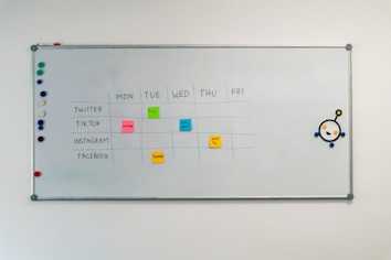 A whiteboard with a weekly social media content schedule is displayed, featuring days of the week across the top and various social media platforms listed on the side. Colorful sticky notes indicate content themes like 'culture', 'fun fact', 'product', and 'how-to'. There are magnets on the left and an eye-catching illustration on the right.