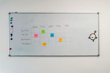 A whiteboard with a weekly social media content schedule is displayed, featuring days of the week across the top and various social media platforms listed on the side. Colorful sticky notes indicate content themes like 'culture', 'fun fact', 'product', and 'how-to'. There are magnets on the left and an eye-catching illustration on the right.