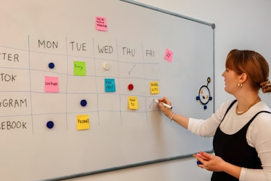 A woman stands beside a whiteboard that serves as a weekly planner, organized with days of the week horizontally and different social media platforms vertically. The board is marked with different colored sticky notes labeled with tasks like 'Culture', 'Funny', 'Fun Fact', 'Diversity', and 'Product'. She uses a pen to make a check mark while looking at the board. The layout is colorful and orderly, indicating a planned schedule for social media marketing.