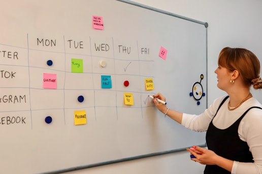 A woman stands beside a whiteboard that serves as a weekly planner, organized with days of the week horizontally and different social media platforms vertically. The board is marked with different colored sticky notes labeled with tasks like 'Culture', 'Funny', 'Fun Fact', 'Diversity', and 'Product'. She uses a pen to make a check mark while looking at the board. The layout is colorful and orderly, indicating a planned schedule for social media marketing.