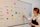 A woman stands beside a whiteboard that serves as a weekly planner, organized with days of the week horizontally and different social media platforms vertically. The board is marked with different colored sticky notes labeled with tasks like 'Culture', 'Funny', 'Fun Fact', 'Diversity', and 'Product'. She uses a pen to make a check mark while looking at the board. The layout is colorful and orderly, indicating a planned schedule for social media marketing.