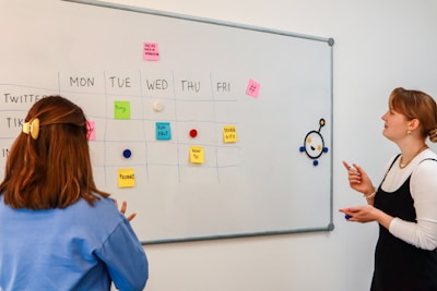 two women standing in front of a whiteboard with sticky notes on it