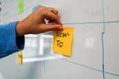 A hand places a yellow sticky note with the words 'How-To' written on it onto a whiteboard. The whiteboard features a grid pattern, and there are other colorful sticky notes attached.