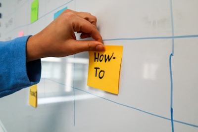A hand places a yellow sticky note with the words 'How-To' written on it onto a whiteboard. The whiteboard features a grid pattern, and there are other colorful sticky notes attached.