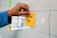 A hand places a yellow sticky note with the words 'How-To' written on it onto a whiteboard. The whiteboard features a grid pattern, and there are other colorful sticky notes attached.