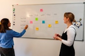 Two people are standing in front of a whiteboard that has a weekly schedule for social media content. The board is divided into sections for Twitter, TikTok, Instagram, and Facebook, with sticky notes indicating different content themes for each day of the week. One person is pointing at the board, while the other holds a red marker.