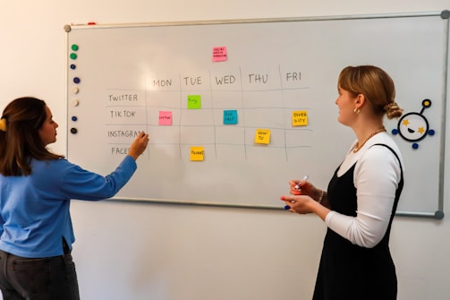 Two people are standing in front of a whiteboard that has a weekly schedule for social media content. The board is divided into sections for Twitter, TikTok, Instagram, and Facebook, with sticky notes indicating different content themes for each day of the week. One person is pointing at the board, while the other holds a red marker.