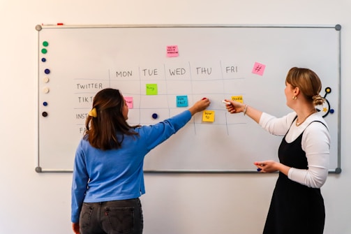 two women writing on a white board with sticky notes