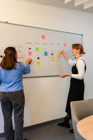 two women standing in front of a whiteboard with sticky notes on it