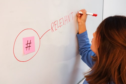 A person is writing the word 'RELEVANT' in red marker on a whiteboard. There is a pink sticky note with a hashtag symbol next to the word, circled in red marker.