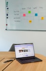 A workspace featuring a whiteboard with a social media content schedule, organized by day and platform, using sticky notes in varying colors. A wooden table holds an open laptop displaying a social media management application, alongside pens and small circular items.