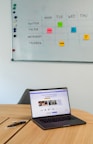 A workspace featuring a whiteboard with a social media content schedule, organized by day and platform, using sticky notes in varying colors. A wooden table holds an open laptop displaying a social media management application, alongside pens and small circular items.