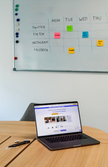 A workspace featuring a whiteboard with a social media content schedule, organized by day and platform, using sticky notes in varying colors. A wooden table holds an open laptop displaying a social media management application, alongside pens and small circular items.