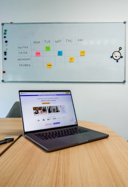 A laptop sits on a wooden table in a room with a whiteboard on the wall. The whiteboard displays a social media content schedule with colored sticky notes under days of the week, planning posts for platforms like Twitter, TikTok, Instagram, and Facebook. The laptop screen shows a website related to social wall solutions.