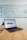 A laptop sits on a wooden table in a room with a whiteboard on the wall. The whiteboard displays a social media content schedule with colored sticky notes under days of the week, planning posts for platforms like Twitter, TikTok, Instagram, and Facebook. The laptop screen shows a website related to social wall solutions.