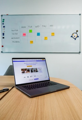A laptop sits on a wooden table in a room with a whiteboard on the wall. The whiteboard displays a social media content schedule with colored sticky notes under days of the week, planning posts for platforms like Twitter, TikTok, Instagram, and Facebook. The laptop screen shows a website related to social wall solutions.