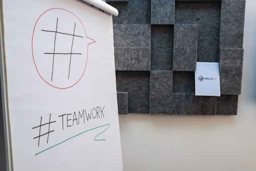 A flip chart displays a simple grid drawing within a red speech bubble and the word '#TEAMWORK' is written below with a green underline. In the background, a textured wall with a modular design holds a small paper labeled 'WALLS.IO'.