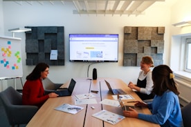 Three people are seated around a rectangular conference table, each with a laptop, engaged in a meeting. A large screen on the wall displays a website or presentation. A whiteboard with colorful sticky notes is visible on the left. Acoustic panels adorn the walls, and the room is well-lit with natural light coming through a window.