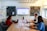 Three people are seated around a rectangular conference table, each with a laptop, engaged in a meeting. A large screen on the wall displays a website or presentation. A whiteboard with colorful sticky notes is visible on the left. Acoustic panels adorn the walls, and the room is well-lit with natural light coming through a window.