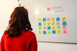 A person with long hair wearing a red sweater is observing a whiteboard. On the whiteboard, there is an arrangement of multi-colored sticky notes with various words and phrases related to company values, such as 'equity', 'teamwork', 'respect', and 'diversity'. The words '# COMPANY VALUES' are prominently written at the top.