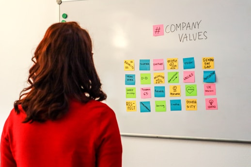 A person with long hair wearing a red sweater is observing a whiteboard. On the whiteboard, there is an arrangement of multi-colored sticky notes with various words and phrases related to company values, such as 'equity', 'teamwork', 'respect', and 'diversity'. The words '# COMPANY VALUES' are prominently written at the top.