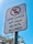 A signpost with a white background and a red circle and line crossing out the silhouette of a dog and cat, indicating that pets are not allowed. The sign contains Arabic text above and below an English message saying 'NO PETS ALLOWED.' The background features a clear blue sky and blurred tree branches.