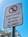 A signpost with a white background and a red circle and line crossing out the silhouette of a dog and cat, indicating that pets are not allowed. The sign contains Arabic text above and below an English message saying 'NO PETS ALLOWED.' The background features a clear blue sky and blurred tree branches.