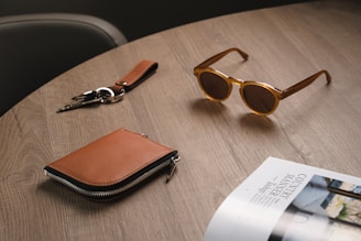 A stylish face cap resting on a polished wooden table beside a pair of sunglasses and a leather wallet.