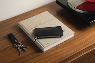 A wooden table holds a beige book titled 'The Monocle Guide to Good Business,' accompanied by a black wallet on top. Next to the book is a keychain with keys and a leather strap. In the background, part of a turntable is visible with a vinyl record on it.