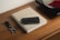 A wooden table holds a beige book titled 'The Monocle Guide to Good Business,' accompanied by a black wallet on top. Next to the book is a keychain with keys and a leather strap. In the background, part of a turntable is visible with a vinyl record on it.