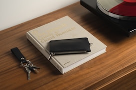 A wooden table holds a beige book titled 'The Monocle Guide to Good Business,' accompanied by a black wallet on top. Next to the book is a keychain with keys and a leather strap. In the background, part of a turntable is visible with a vinyl record on it.